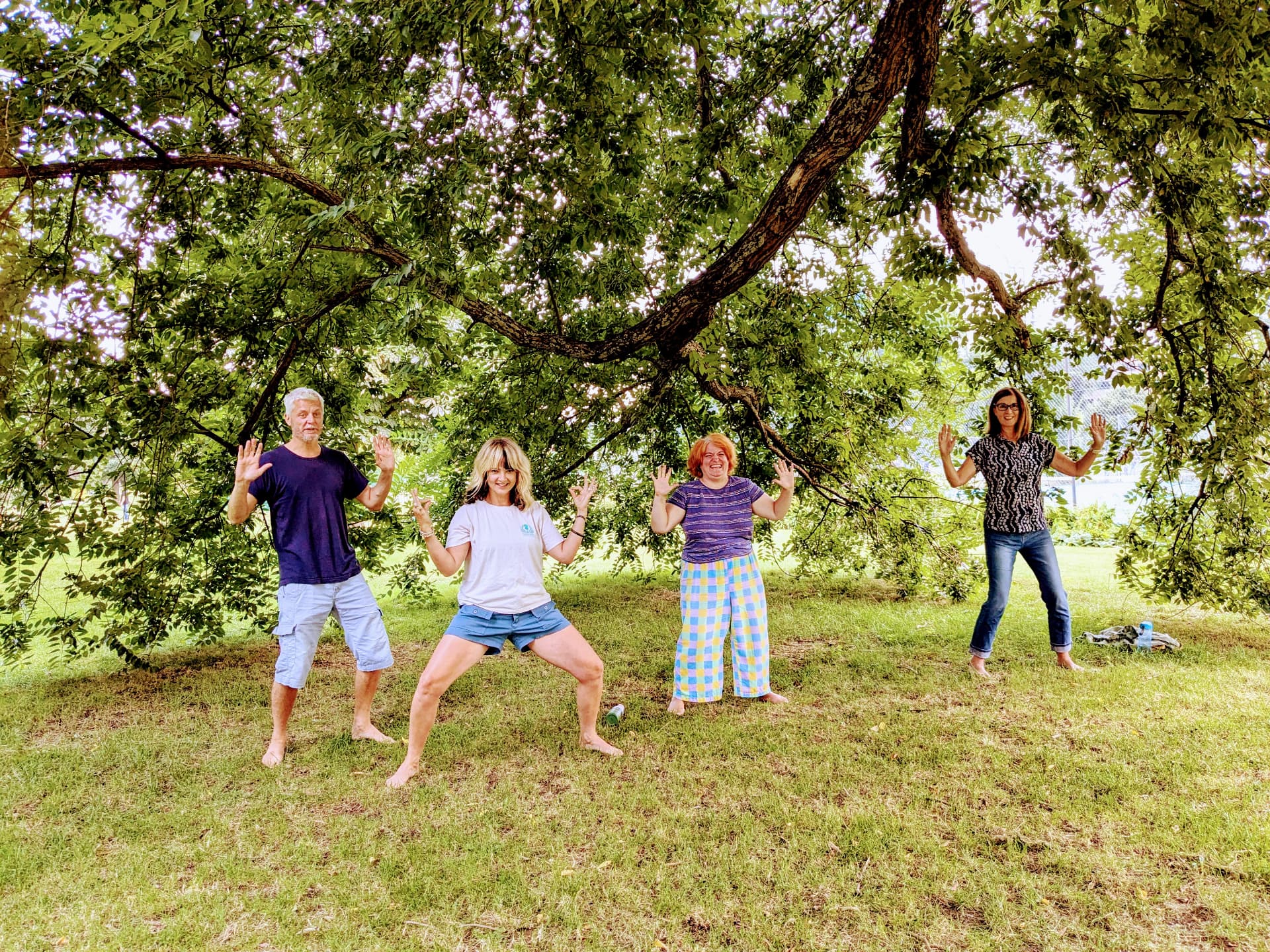 Four smiling qigong students practise together under a leafy tree after a class